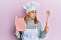 Hispanic young chef woman reading recipes book smiling looking to the side and staring away thinking Royalty Free Stock Photo