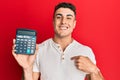 Hispanic young man showing calculator device pointing finger to one self smiling happy and proud