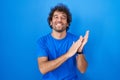 Hispanic young man standing over blue background clapping and applauding happy and joyful, smiling proud hands together Royalty Free Stock Photo
