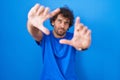 Hispanic young man standing over blue background doing frame using hands palms and fingers, camera perspective Royalty Free Stock Photo