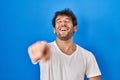 Hispanic young man standing over blue background laughing at you, pointing finger to the camera with hand over body, shame Royalty Free Stock Photo