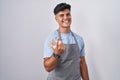 Hispanic young man wearing apron over white background beckoning come here gesture with hand inviting welcoming happy and smiling Royalty Free Stock Photo