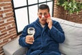 Hispanic young man wearing bathrobe and eye bags patches drinking wine speaking on the phone clueless and confused expression Royalty Free Stock Photo