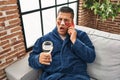 Hispanic young man wearing bathrobe and eye bags patches drinking wine speaking on the phone in shock face, looking skeptical and Royalty Free Stock Photo