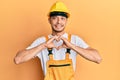 Hispanic young man wearing builder uniform and safety hardhat smiling in love doing heart symbol shape with hands