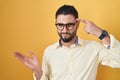 Hispanic young man wearing business clothes and glasses confused and annoyed with open palm showing copy space and pointing finger