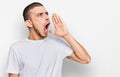 Hispanic young man wearing casual white t shirt shouting and screaming loud to side with hand on mouth