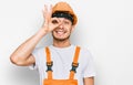 Hispanic young man wearing handyman uniform and safety hardhat smiling happy doing ok sign with hand on eye looking through Royalty Free Stock Photo