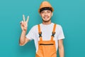 Hispanic young man wearing handyman uniform and safety hardhat smiling with happy face winking at the camera doing victory sign Royalty Free Stock Photo