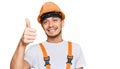 Hispanic young man wearing handyman uniform and safety hardhat smiling happy and positive, thumb up doing excellent and approval Royalty Free Stock Photo
