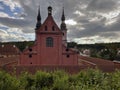 Historic church with red facade and three ornate spires, surrounded by greenery and rooftops, under a dramatic cloudy sky with Royalty Free Stock Photo