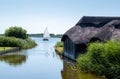 Historic thatched boathouses amongst the green reeds on Hickling Broad, east of Norwich, in Norfolk UK. Sailing boat in the dist