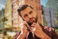 Hm... Portrait of thoughtful bearded man talking by phone and touching his stubble while standing on the street Royalty Free Stock Photo