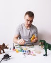 Hobby: a man paints dinosaur toy figures with a brush and paints on a white table