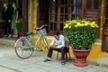 Hoi Local man with smartphone is sitting on a plastic chair near to yellow house with flower pots with yellow chrysanthemums.