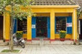 Yellow house with blue doors and windows and flower pots with yellow chrysanthemums and motorbike next to it. Small book shop.