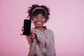 Holding black phone in hand. Attractive afro american woman in casual clothes at pink background in the studio