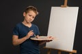 Holding a palette with paints little boy in blue t-shirt looking sad standing next to the easel with canvas on it