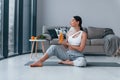 Holds glass with orange juice in hands. Young woman with slim body shape in sportswear have fitness day indoors at home Royalty Free Stock Photo