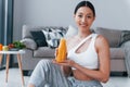 Holds glass with orange juice in hands. Young woman with slim body shape in sportswear have fitness day indoors at home Royalty Free Stock Photo