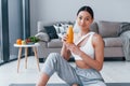 Holds glass with orange juice in hands. Young woman with slim body shape in sportswear have fitness day indoors at home Royalty Free Stock Photo