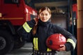 Holds red hat in hands. Female firefighter in protective uniform standing near truck