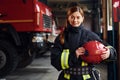 Holds red hat in hands. Female firefighter in protective uniform standing near truck