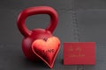 Holiday fitness, red kettlebell, with a metal red heart, on a black gym floor, with red valentine card