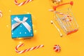 Holiday shopping. A toy shopping cart and a gift box. Yellow background with candys and christmas ball. Close up. Top view. Royalty Free Stock Photo