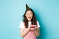 Holidays and celebration. Cheerful birthday girl in party hat holding bday cake and smiling, making wish on lit candle