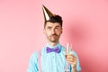 Holidays and celebration concept. Grumpy guy wearing birthday party hat and holding glass of champagne, looking up with