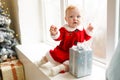 Holidays and childhood concept - sweet baby girl in santa costume sitting with xmas gift box at the window near christmas tree