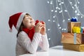 Holidays and Christmas Ideas. Dreaming Winsome Caucasian Female Girl in Santa Hat and White Shirt Holding Tiny Red Gift Box Near