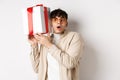 Holidays concept. Funny young man in glasses shaking gift box, listening what inside present and gasping amazed Royalty Free Stock Photo