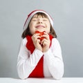 Holidays Concepts. One Dreaming Winsome Caucasian Female Kid in Santa Hat and White Shirt Holding Tiny Red Gift Box in Front of