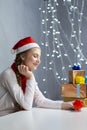 Holidays Ideas. Dreaming Winsome Caucasian Female Girl in Santa Hat and White Shirt Holding Tiny Red Gift Box Near Stack of