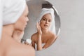 Home beauty routine. Young woman applying moisturising cream on face looking at miror in bathroom, free space