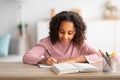 Home education concept. Cute african american schoolgirl doing homework and smiling, sitting at table with book Royalty Free Stock Photo