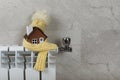 Home heating radiator. Model of a house wrapped in a scarf and hat on a radiator indoors against a gray wall. Royalty Free Stock Photo
