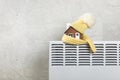 Home heating radiator. Model of a house wrapped in a scarf and hat on a radiator indoors against a gray wall. Royalty Free Stock Photo