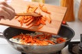 Home kitchen. A woman with a wooden cutting board adds carrots to a hot frying pan with vegetable oil and onions. Close-up