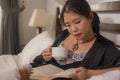 Home lifestyle morning portrait of young beautiful and happy Asian Chinese woman in nightgown reading a book in bed enjoying the Royalty Free Stock Photo