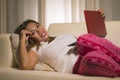 Young beautiful and natural Asian Indonesian woman in pajamas pants relaxed on couch networking using laptop computer shopping Royalty Free Stock Photo