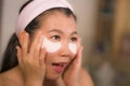 Home lifestyle portrait of young happy and beautiful Asian Korean woman at bathroom mirror applying panda eye patch anti-aging