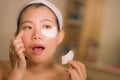 Home lifestyle portrait of young happy and beautiful Asian Korean woman at bathroom mirror applying panda eye patch anti-aging