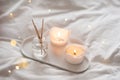 Home liquid fragrance in glass bottle and burning candles staying on white ceramic tray in bed close up. Royalty Free Stock Photo