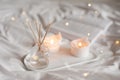 Home liquid fragrance in glass bottle and burning candles staying on white ceramic tray in bed close up. Royalty Free Stock Photo