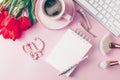 Home office workplace: cup of coffee, empty notebook, keyboard, tulip flowers, beads, headphones on pink background