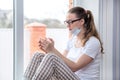 Home quarantine. Caucasian woman on sick leave is sitting at window in a medical protective mask, drinking medicinal herbal tea or Royalty Free Stock Photo