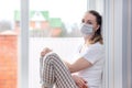 Home quarantine. Caucasian woman on sick leave is sitting at window in a medical protective mask, looking sad. Poor health, a Royalty Free Stock Photo